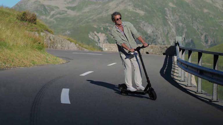 großer Mann mit weisser Hose fährt eine Bergstrasse mit dem eScooter VMAX VX2 hoch